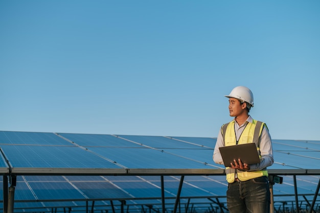 Un joven ingeniero inspector asiático usa una computadora portátil que trabaja en una granja solar Supervisor técnico masculino con casco blanco Comprobación del funcionamiento del panel solar y fotovoltaico en el espacio de copia de la estación