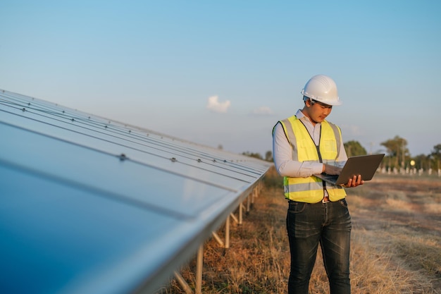 Un joven ingeniero inspector asiático usa una computadora portátil que trabaja en una granja solar Supervisor técnico masculino con casco blanco Comprobación del funcionamiento del panel solar y fotovoltaico en el espacio de copia de la estación