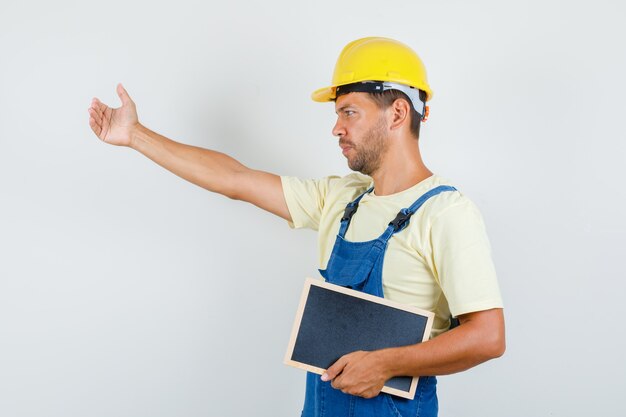 Joven ingeniero invitando a venir y sosteniendo pizarra en uniforme.