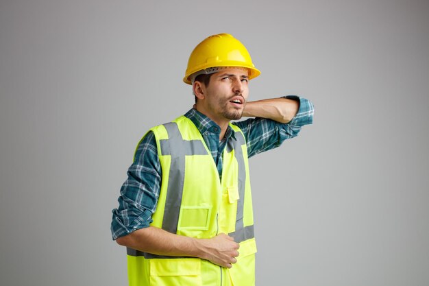 Joven ingeniero malsano con casco de seguridad y uniforme mirando hacia arriba manteniendo las manos en el vientre y detrás del cuello aislado en fondo blanco