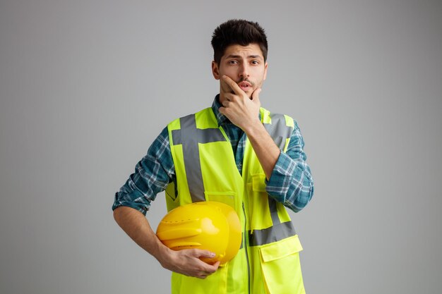 Un joven ingeniero pensativo que usa uniforme con casco de seguridad mirando a la cámara mientras mantiene la mano en la barbilla aislada en el fondo blanco