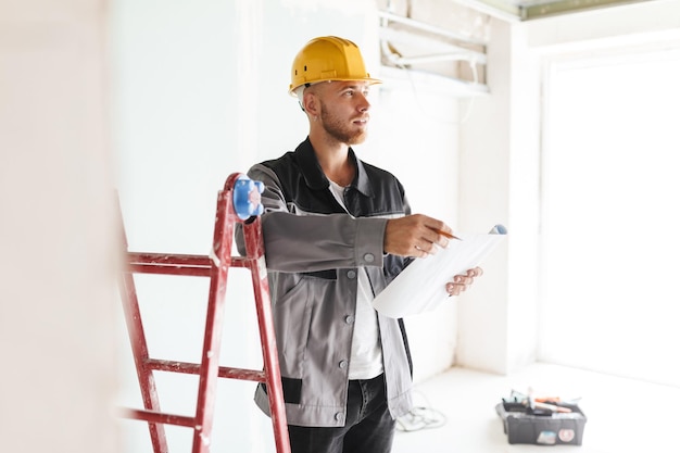 Joven ingeniero en ropa de trabajo y casco amarillo mirando soñadoramente a un lado sosteniendo un plan de nuevos apartamentos en manos con una escalera roja cerca de reparar el piso