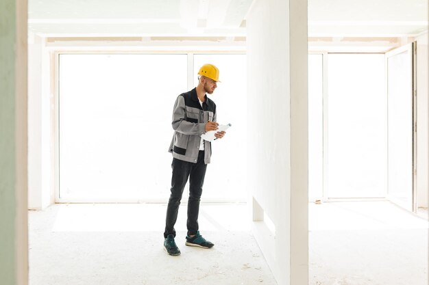 Joven ingeniero en ropa de trabajo y casco amarillo sosteniendo un plan de nuevos apartamentos mirando cuidadosamente en la pared con una gran ventana en el fondo