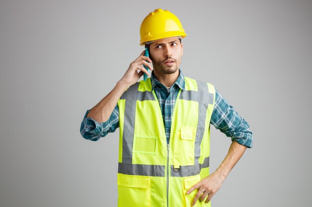 Un joven ingeniero serio con casco de seguridad y uniforme manteniendo la mano en la cintura mirando al costado mientras habla por teléfono aislado de fondo blanco