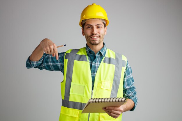 Un joven ingeniero sonriente con casco de seguridad y uniforme sosteniendo un bloc de notas y un lápiz mirando la cámara apuntando hacia abajo aislada en el fondo blanco