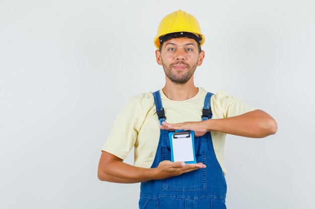 Joven ingeniero sosteniendo un mini portapapeles en uniforme y mirando alegre, vista frontal.