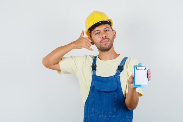 Joven ingeniero sosteniendo portapapeles con gesto de teléfono en uniforme, vista frontal.