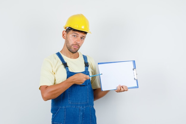 Foto gratuita joven ingeniero en uniforme apuntando al portapapeles, vista frontal.