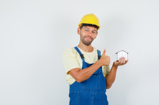 Joven ingeniero en uniforme sosteniendo el modelo de casa con el pulgar hacia arriba y mirando alegre, vista frontal.