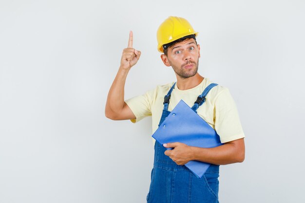 Joven ingeniero en uniforme sosteniendo el portapapeles y apuntando hacia arriba, vista frontal.