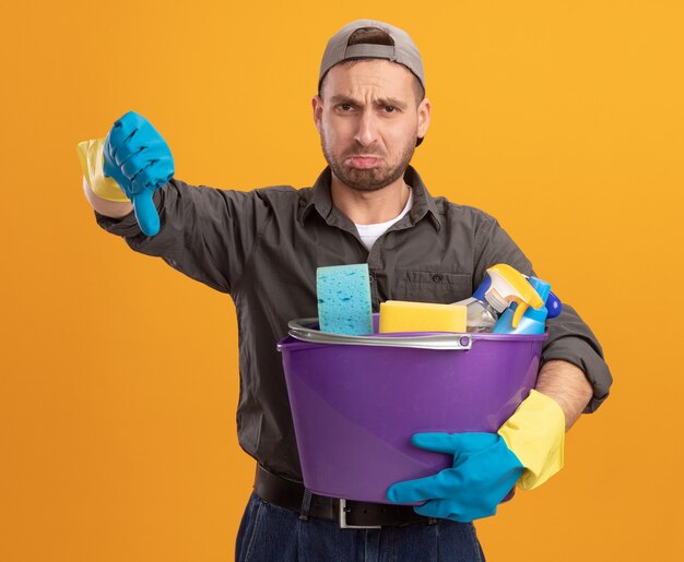 Joven insatisfecho con ropa casual y gorra en guantes de goma sosteniendo un balde con herramientas de limpieza mostrando los pulgares hacia abajo de pie sobre la pared naranja