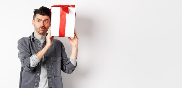 Foto gratuita joven intrigado mirando confundido a la cámara tratando de adivinar qué hay dentro de la caja de regalo del amante shaki