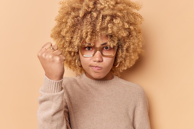 Una joven irritada con el pelo rizado aprieta el puño dice que les mostraré los labios de los bolsos se siente indignada usa anteojos saltadores casuales aislados sobre el fondo marrón del estudio parece enojada Los odio a todos