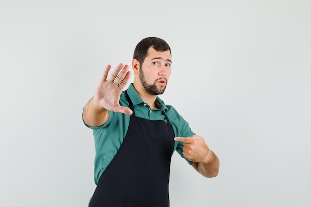 Joven jardinero apuntando a sí mismo mientras muestra gesto de parada en camiseta, vista frontal del delantal.