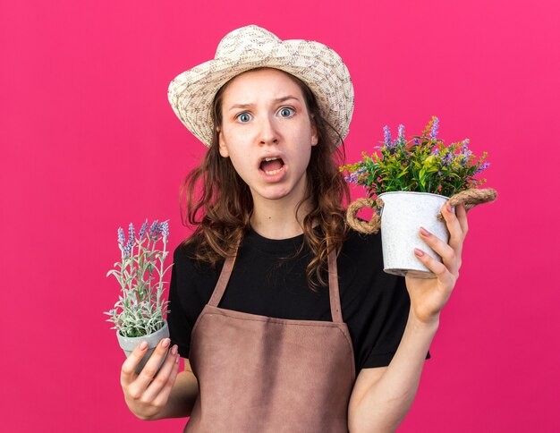 Joven jardinero asustado vistiendo sombrero de jardinería sosteniendo flores en macetas