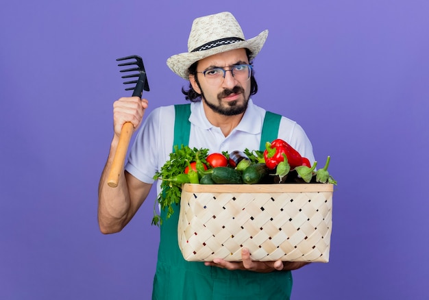 Joven jardinero barbudo hombre vestido con mono y sombrero con caja llena de verduras y mini rastrillo mirando al frente con cara seria de pie sobre la pared azul