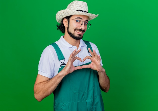 Joven jardinero barbudo hombre vestido con mono y sombrero haciendo gesto de corazón con los dedos sonriendo con cara feliz