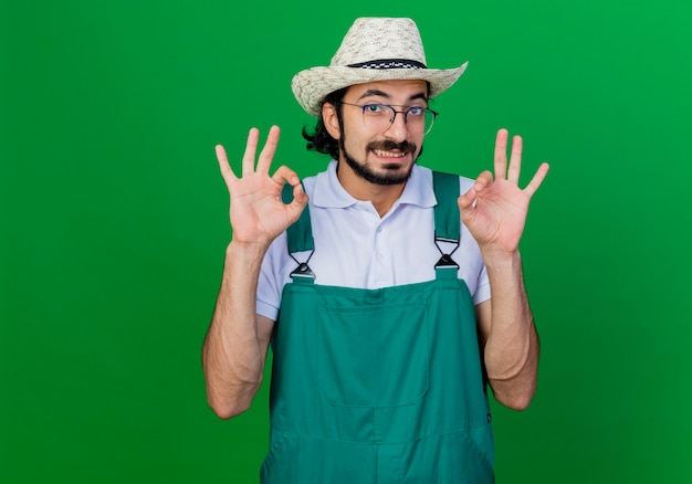 Joven jardinero barbudo hombre vestido con mono y sombrero sonriendo mostrando signo ok