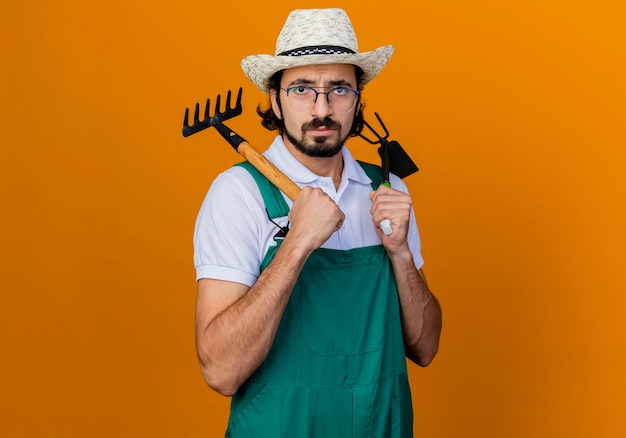 Joven jardinero barbudo hombre vestido con mono y sombrero sosteniendo azadón y mini rastrillo mirando al frente con cara seria de pie sobre la pared naranja