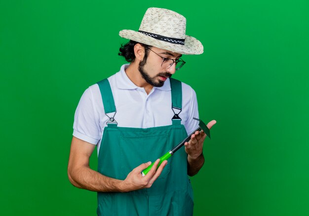 Joven jardinero barbudo hombre vestido con mono y sombrero sosteniendo azadón mirándolo con cara seria