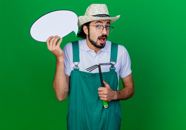 Joven jardinero barbudo hombre vestido con mono y sombrero sosteniendo azadón y signo de burbuja de discurso en blanco sorprendido