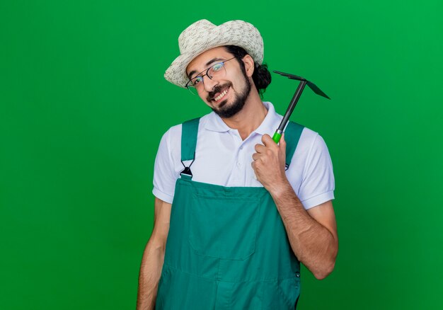 Joven jardinero barbudo hombre vestido con mono y sombrero sosteniendo azadón sonriendo alegremente