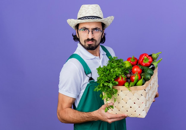 Foto gratuita joven jardinero barbudo hombre vestido con mono y sombrero sosteniendo una caja llena de verduras mirando al frente con el ceño fruncido serio de pie sobre la pared azul