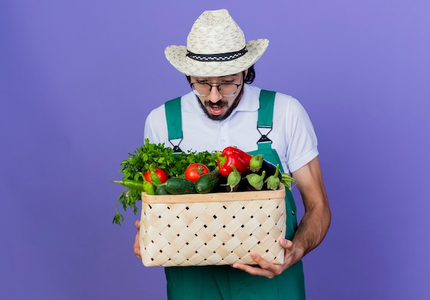 Joven jardinero barbudo hombre vestido con mono y sombrero sosteniendo una caja llena de verduras mirándolo sorprendido y asombrado de pie sobre la pared azul