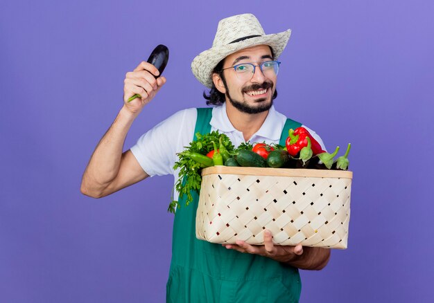 Joven jardinero barbudo hombre vestido con mono y sombrero sosteniendo una caja llena de verduras mostrando berenjena sonriendo alegremente de pie sobre la pared azul