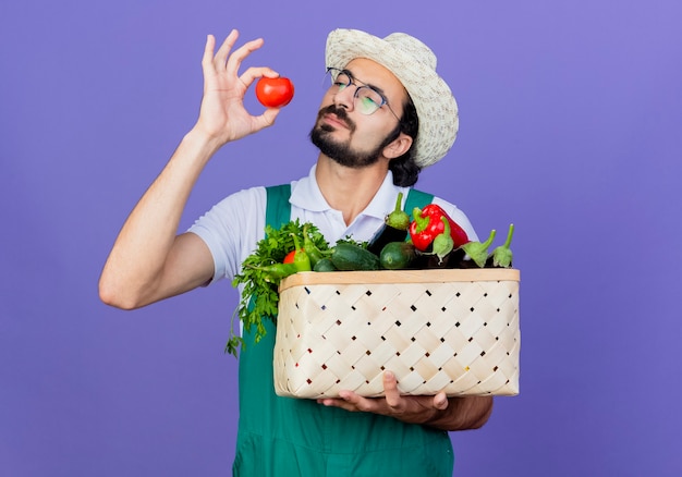 Joven jardinero barbudo hombre vestido con mono y sombrero sosteniendo una caja llena de verduras y tomate fresco mirándolo intrigado de pie sobre la pared azul