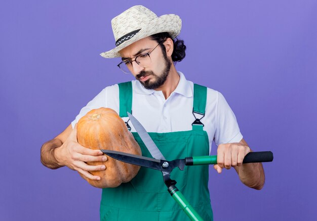 Joven jardinero barbudo hombre vestido con mono y sombrero sosteniendo calabaza mirándolo con cara seria de pie sobre la pared azul