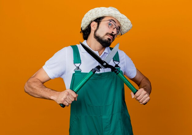 Joven jardinero barbudo hombre vestido con mono y sombrero sosteniendo cortasetos mirando confundido y desconcertado de pie