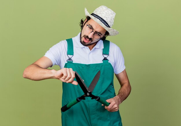 Joven jardinero barbudo hombre vestido con mono y sombrero sosteniendo cortasetos mirándolo desconcertado parado sobre la pared verde claro