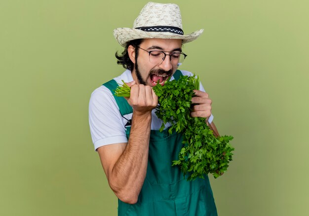 Joven jardinero barbudo hombre vestido con mono y sombrero sosteniendo hierbas frescas comiéndolo de pie sobre la pared verde claro