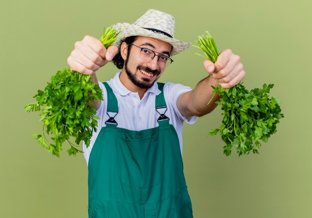 Joven jardinero barbudo hombre vestido con mono y sombrero sosteniendo hierbas frescas lookign a cámara sonriendo con cara feliz