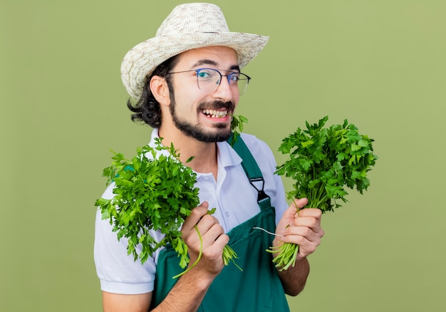 Joven jardinero barbudo hombre vestido con mono y sombrero sosteniendo hierbas frescas mirando al frente sonriendo con cara feliz de pie sobre la pared verde claro