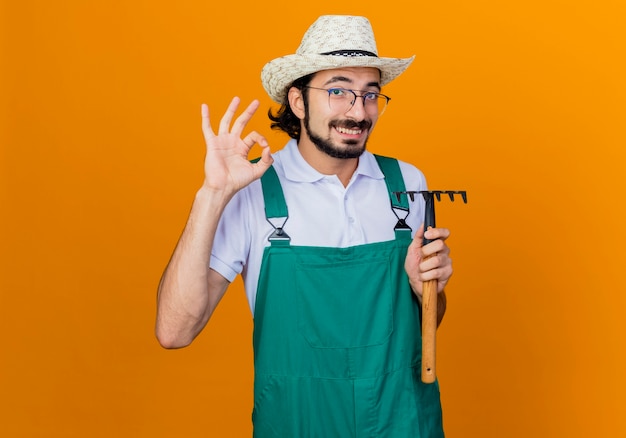 Joven jardinero barbudo hombre vestido con mono y sombrero sosteniendo mini rastrillo mirando al frente sonriendo mostrando signo ok de pie sobre la pared naranja