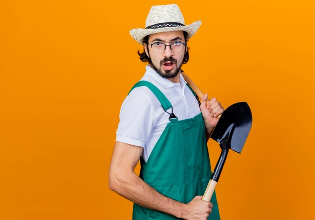 Joven jardinero barbudo hombre vestido con mono y sombrero sosteniendo pala mirando al frente confundido parado sobre pared naranja