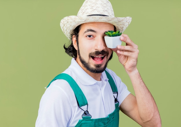 Joven jardinero barbudo hombre vestido con mono y sombrero sosteniendo planta en maceta mirando al frente sonriendo de pie sobre la pared verde claro