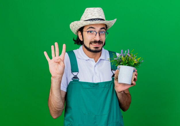 Joven jardinero barbudo hombre vestido con mono y sombrero sosteniendo planta en maceta mostrando el número cuatro