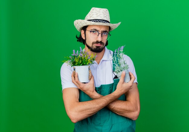 Joven jardinero barbudo hombre vestido con mono y sombrero sosteniendo plantas en macetas con cara de seriou disgustado