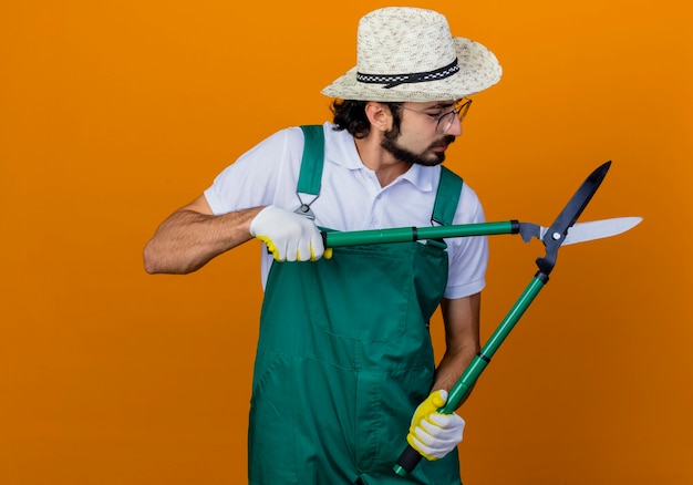 Joven jardinero barbudo hombre vestido con mono y sombrero sosteniendo podadoras de setos mirando podadoras con cara seria