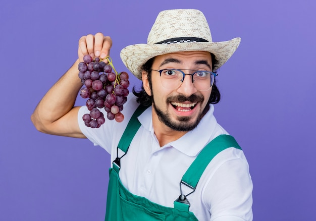 Joven jardinero barbudo hombre vestido con mono y sombrero sosteniendo racimo de uva mirando al frente sonriendo de pie sobre la pared azul