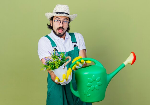 Joven jardinero barbudo hombre vestido con mono y sombrero sosteniendo regadera y planta en maceta mirando al frente confundido parado sobre pared verde claro