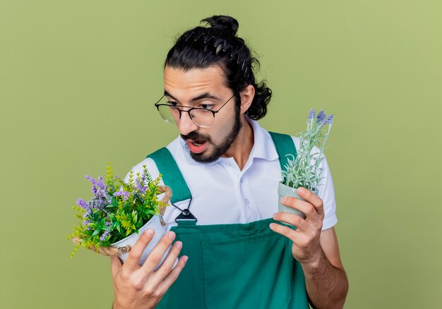 Joven jardinero barbudo hombre vestido con mono sosteniendo plantas en macetas mirándolas confundidas tratando de tomar una decisión de pie sobre la pared verde claro