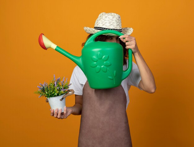 Joven jardinero complacido en uniforme vistiendo sombrero de jardinería sosteniendo flor en maceta con la cara cubierta con regadera