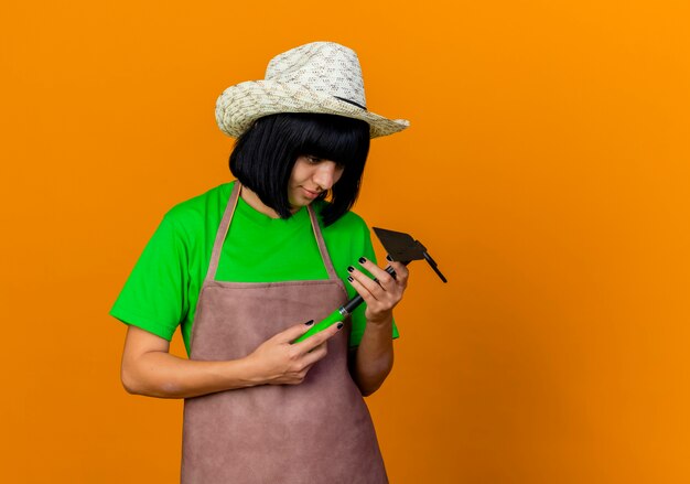 Joven jardinero complacido en uniforme vistiendo sombrero de jardinería sostiene y mira azada rastrillo