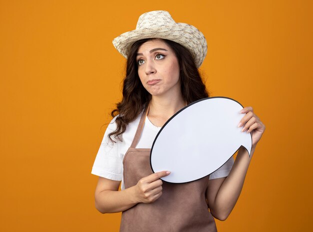 Foto gratuita joven jardinero decepcionado en uniforme con sombrero de jardinería sostiene bocadillo mirando al lado aislado en la pared naranja