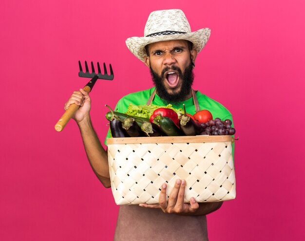 Joven jardinero enojado chico afroamericano con sombrero de jardinería sosteniendo canasta de verduras con rastrillo