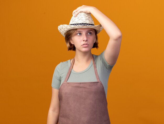 Joven jardinero eslavo confiado con sombrero de jardinería pone la mano en el sombrero mirando al lado en naranja